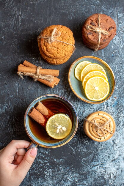 Vista vertical de limones frescos y mano sosteniendo una taza de té negro con galletas de canela apiladas sobre fondo oscuro