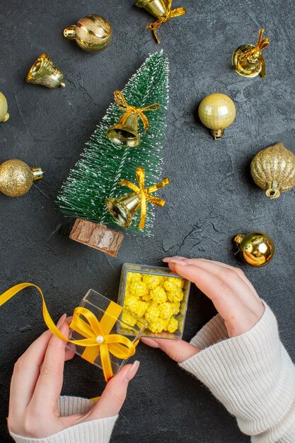 Vista vertical de la mano que sostiene cajas de regalo y accesorios de decoración del árbol de navidad sobre fondo oscuro