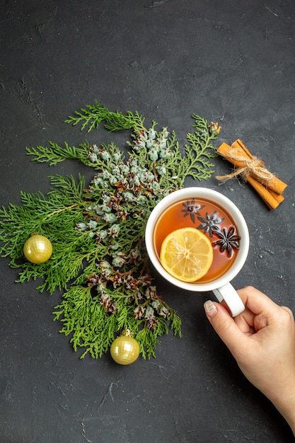 Vista vertical de la mano sosteniendo una taza de té negro accesorios navideños y limones canela sobre fondo negro