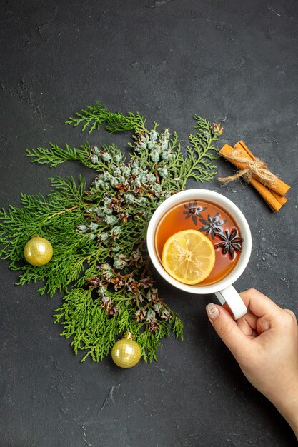 Vista vertical de la mano sosteniendo una taza de té negro accesorios navideños y limones canela sobre fondo negro