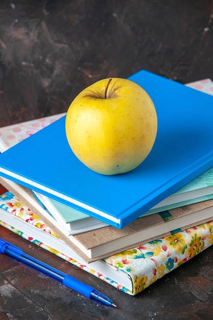 Vista vertical de manzana amarilla fresca en cuadernos apilados y bolígrafo en el lado izquierdo sobre fondo oscuro