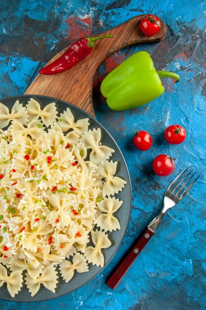 Vista vertical de pasta farfalle italiana cruda con verduras en una placa negra sobre tabla de cortar de madera y tenedor junto a pimientos tomates sobre fondo azul.