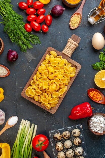 Vista vertical de pastas crudas en una tabla de cortar marrón huevos verduras diversas especias en la oscuridad