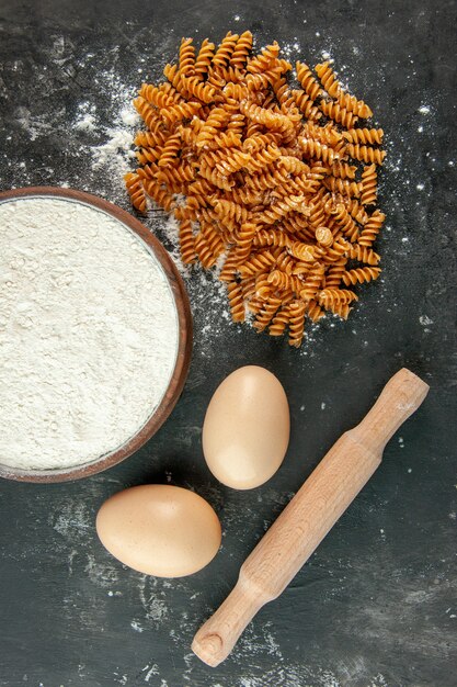 Vista vertical de pastas italianas crudas dos huevos y harina en un cuenco marrón con rodillo sobre fondo gris