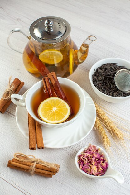 Vista vertical de la pausa para el té con canela, lima y limón en una taza y una olla alrededor de los ingredientes necesarios sobre fondo blanco.