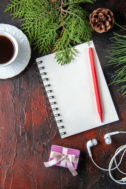 Vista vertical de ramas de abeto, una taza de té negro, accesorios de decoración, auriculares blancos y regalo junto al cuaderno con bolígrafo sobre fondo oscuro