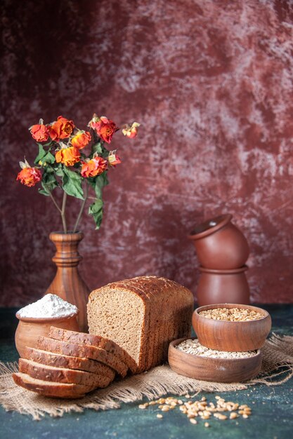 Vista vertical de rebanadas de pan negro harina en un cuenco y avena cruda de trigo sobre una toalla de color nude y macetas sobre fondo de colores mezclados