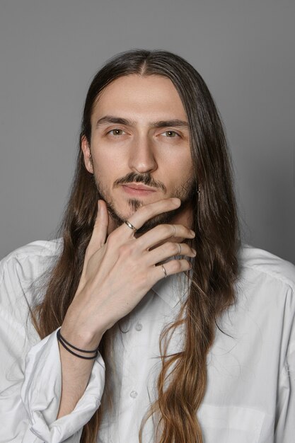 Vista vertical recortada de un joven barbudo pensativo pensativo con cabello largo con accesorios elegantes y camisa blanca, frotándose la barbilla mientras piensa en algo, con expresión incierta
