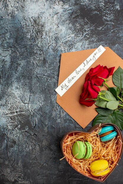 Vista vertical de rosas rojas y sobre con carta de amor y diferentes macarons sobre fondo oscuro helado