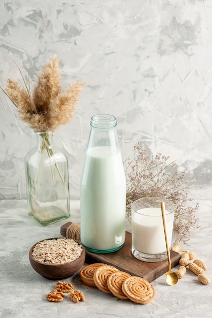 Vista vertical de la taza de la botella de vidrio abierta llena de leche en una cuchara de tabla de cortar de madera y avena de nuez en galletas de olla marrón en la pared gris
