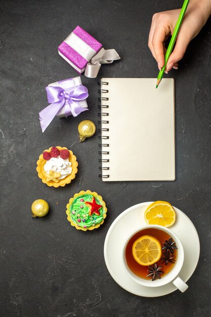 Vista vertical de una taza de té negro con limón servido con galletas y regalos junto al cuaderno con lápiz sobre fondo oscuro