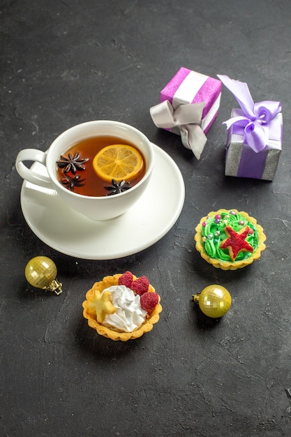 Vista vertical de una taza de té negro con limón servido con galletas y regalos sobre fondo oscuro