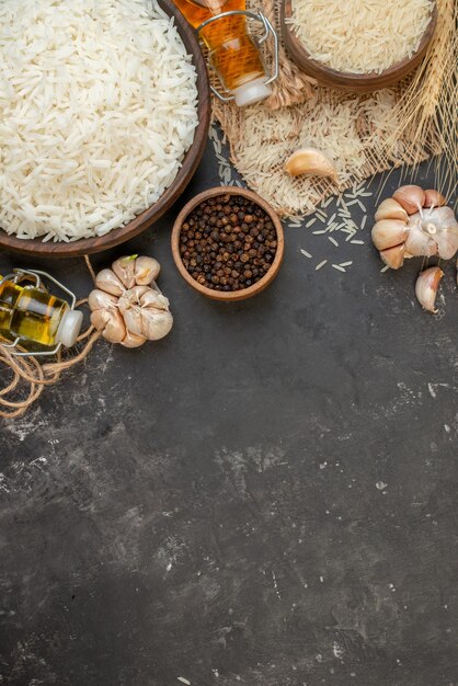 Vista vertical de tazones largos perfectos de arroz marrón sobre una toalla de color desnudo, botellas de aceite caídas, ajos, pimientos en la parte superior sobre fondo oscuro