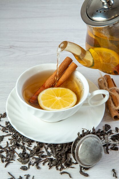 Vista vertical de té de hierbas que se vierte de la olla en una taza blanca y limones de canela alrededor de té seco sobre fondo blanco.