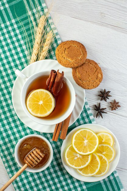 Vista vertical de té negro en una taza blanca con limas canela y limón junto a deliciosas galletas de miel en una toalla despojada verde sobre fondo blanco.