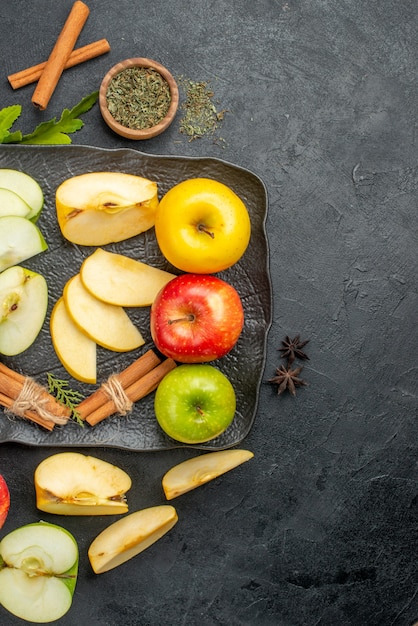 Vista vertical de verde amarillo y rojo en rodajas y manzanas frescas enteras en una bandeja negra y limones canela en el lado derecho sobre un fondo oscuro