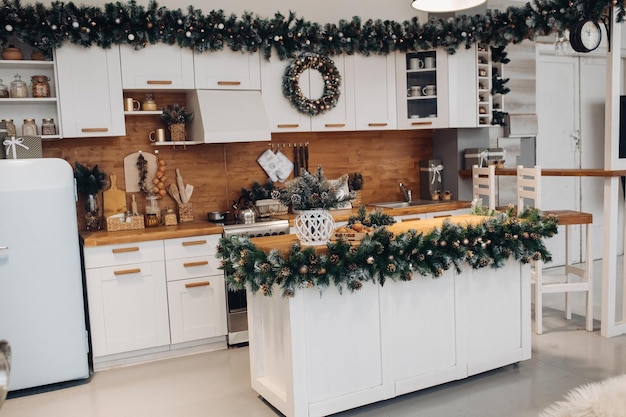Vistas a la cocina moderna con alacenas blancas y mesa marrón decorada con ramas de abeto verde natural y corona de Navidad. Decoraciones de navidad.