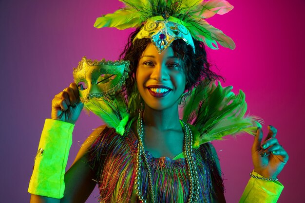 Vistoso. Hermosa mujer joven en carnaval, elegante disfraz de mascarada con plumas bailando en la pared degradada en neón. Concepto de celebración navideña, tiempo festivo, baile, fiesta, diversión.