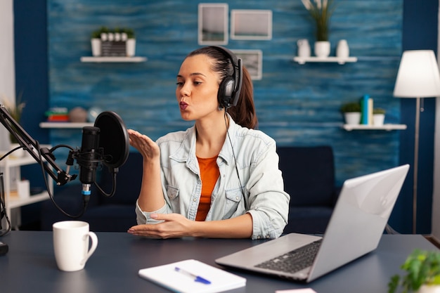 Vlogger dando un beso volador mientras hace un video blog diario en línea. Programa de entrevistas de grabación de influcencer digital en home studio brodcast con auriculares, micrófono de podcast profesional y computadora portátil moderna