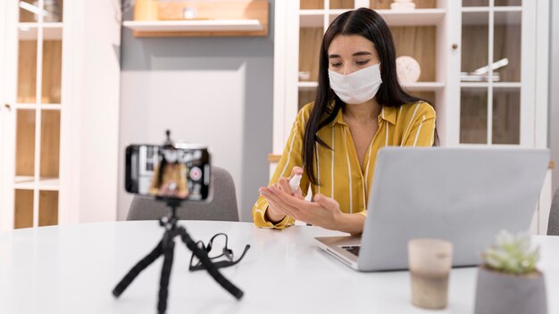 Vlogger femenina en casa con máscara médica y smartphone