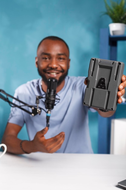Vlogger sonriente presentando la batería de la cámara, revisando el equipo de videografía mientras graba video. Blogger que promociona equipo de fotografía mientras transmite en vivo en el canal de redes sociales