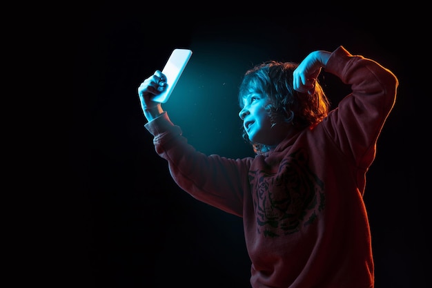 Vlogging con smartphone. Retrato de niño caucásico sobre fondo oscuro de estudio en luz de neón. Preciosa modelo de pelo rizado.