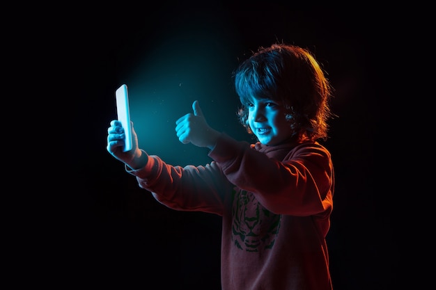 Vlogs con smartphone, pulgar hacia arriba. Retrato de niño caucásico sobre fondo oscuro en luz de neón. Preciosa modelo rizada. Concepto de emociones humanas, expresión facial, ventas, publicidad, tecnología moderna, gadgets.