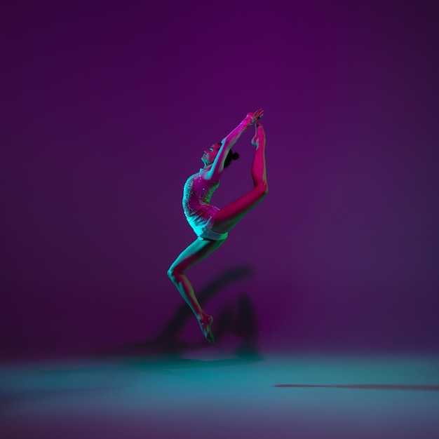 Foto gratuita volador. joven atleta femenina, artista de gimnasia rítmica bailando, entrenando aislada en un estudio morado con luz de neón. hermosa chica practicando con equipo. gracia en el desempeño.