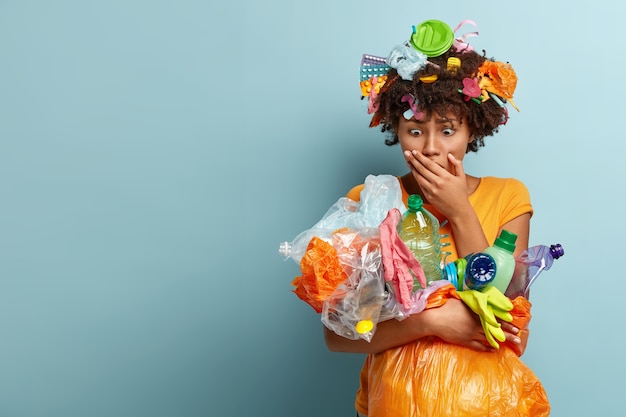 Foto gratuita voluntaria mujer negra aterrorizada mira la basura plástica, demuestra conciencia de la contaminación, viste una camiseta amarilla informal, sostiene basura, se para sobre una pared azul.