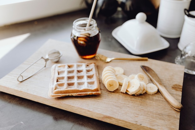 Waffle y rodajas de plátano en una bandeja de madera junto a un tarro de miel