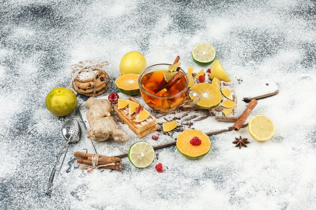 Waffles de vista de alto ángulo y obleas de arroz en la tabla de cortar blanca con té de hierbas, frutas cítricas, canela y colador de té en la superficie de mármol gris oscuro. horizontal