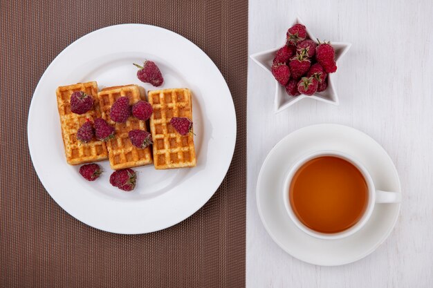 Waffles de vista superior en un plato con frambuesas y una taza de té en una mesa blanca