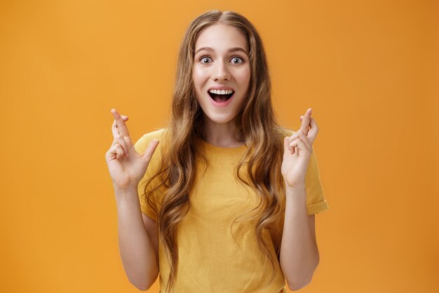 Wais-up shot de una atractiva mujer interesada, divertida y entusiasta con una camiseta casual cruzando los dedos para la buena suerte, sonriendo, asombrada y encantada de estar feliz y con la esperanza de que las cosas vayan bien.