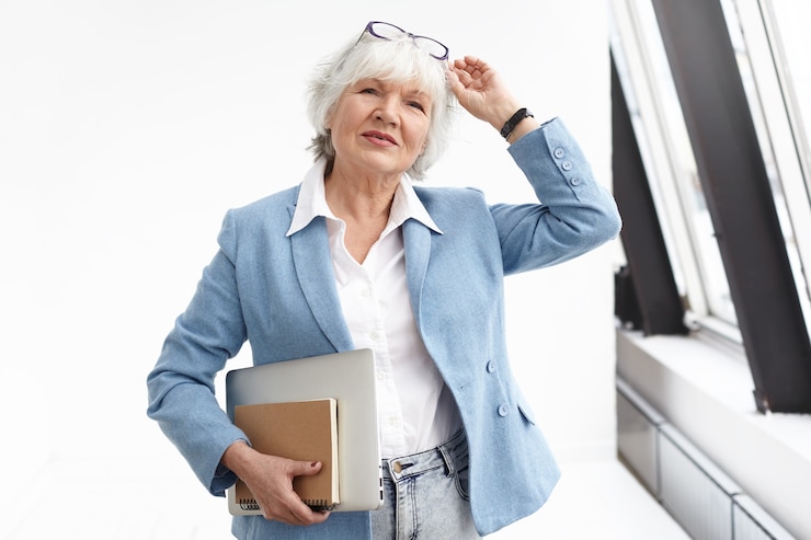 Photo Gratis waist up view of smart classy senior businesswoman wearing stylish blue jacket, jeans and white shirt adjusting glasses on her head, carrying book and laptop, heading to meeting, standing by window