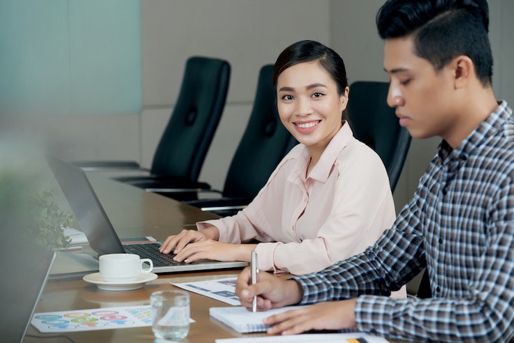 Photo Gratis wanita asia tersenyum duduk di meja pertemuan dengan laptop, dan rekan pria menulis di buku catatan