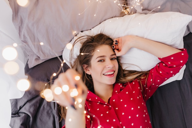 Winsome girl viste pijamas rojos de moda divirtiéndose en la mañana. Preciosa modelo de mujer con hermosa sonrisa en sábana negra y almohada blanca.