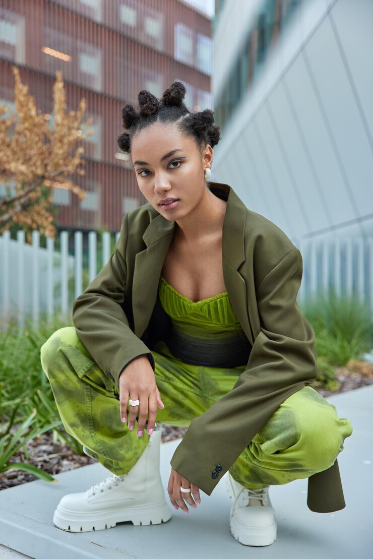 Photo Gratis woman kneels down outside dressed in stylish green apparel white boots wears blue eyeliner has hairstyle looks self assured at camera. urban style and youth concept