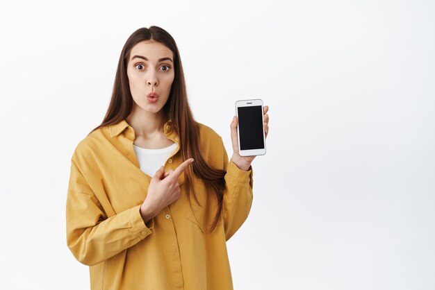 Wow, esto es interesante Mujer joven curiosa que señala la pantalla del teléfono inteligente con cara emocionada y sorprendida encontró una aplicación genial en la tienda web en línea o una aplicación de entrega con fondo blanco