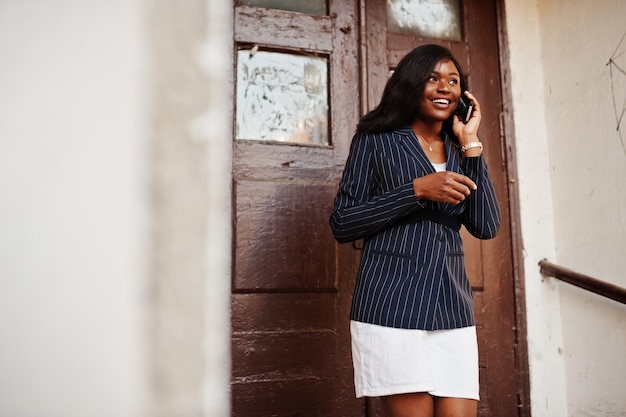 Éxito elegante mujer afroamericana en chaqueta y falda contra la vieja puerta de madera con el teléfono inteligente en las manos