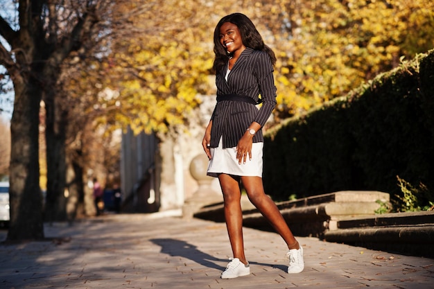 Éxito elegante mujer afroamericana en chaqueta y falda posada en el soleado día de otoño en la calle