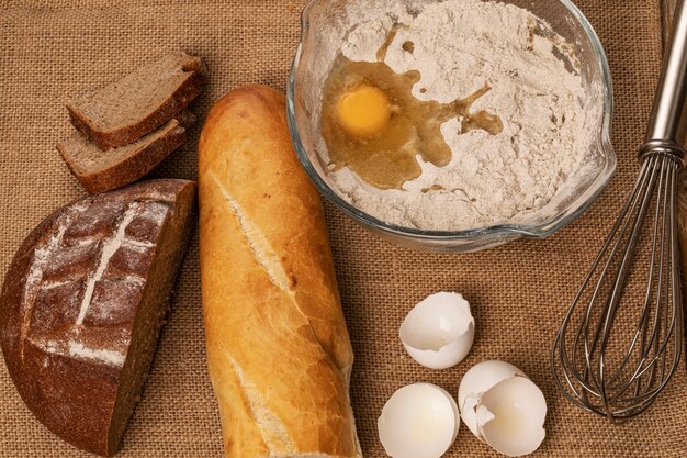 Yema de huevo sobre la harina. Cáscaras de huevo, baguette y rebanadas de pan de centeno y una batidora manual sobre un trozo de arpillera. Vista superior.