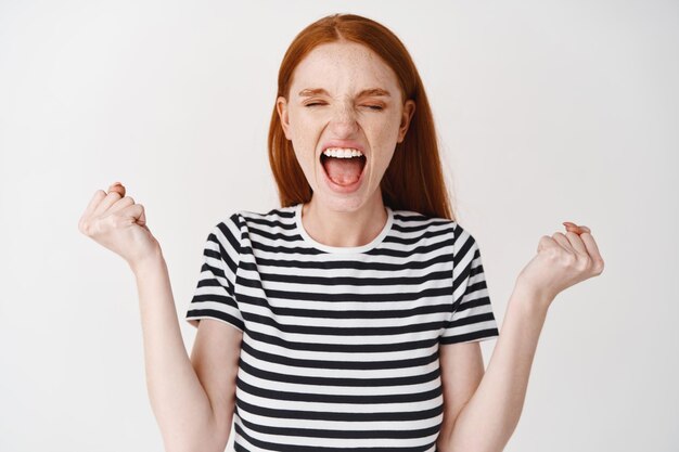 Yo gano. Feliz mujer pelirroja celebra el logro, grita de alegría, aprieta los puños en señal de triunfo, de pie sobre la pared blanca