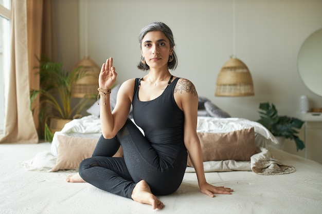 Yogui femenina avanzada joven flexible con canas prematuras sentada en el suelo en postura ardha matsyendrasana, haciendo un giro espinal sentado para mejorar la digestión y aliviar el dolor de espalda