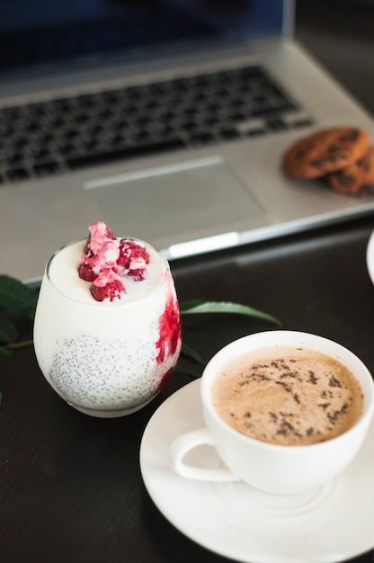 Yogurt con frambuesas; Taza de café y galletas en la computadora portátil contra el fondo negro