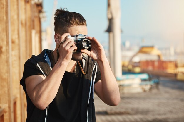 Young enfocó a un chico europeo de pie en el puerto mirando a través de la cámara mientras tomaba fotos de mar o yates, caminando por la ciudad para tomar fotos geniales para la revista. Ángulo de búsqueda de camarógrafo talentoso