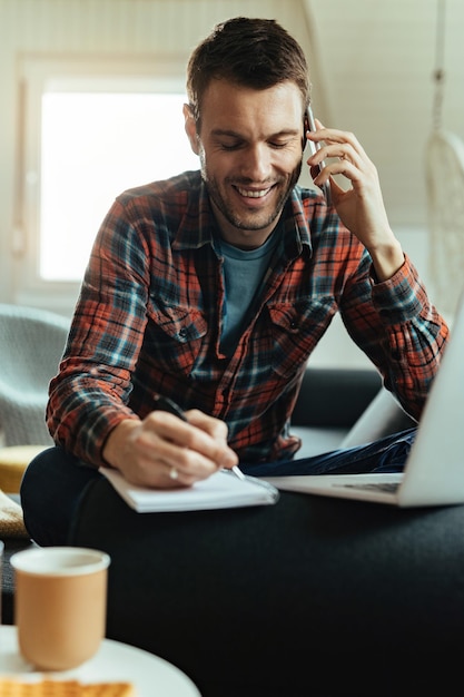 Yung hombre feliz comunicándose por teléfono móvil y escribiendo notas en la sala de estar.