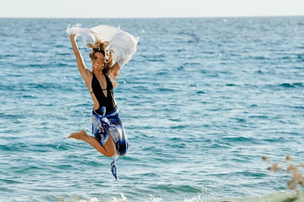 Yung mujer juguetona con un chal saltando en la playa y divirtiéndose en el día de verano