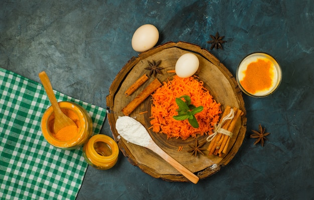 Zanahoria rallada con huevos, harina, especias, leche, paño de picnic sobre tabla de madera y fondo de estuco, vista superior.