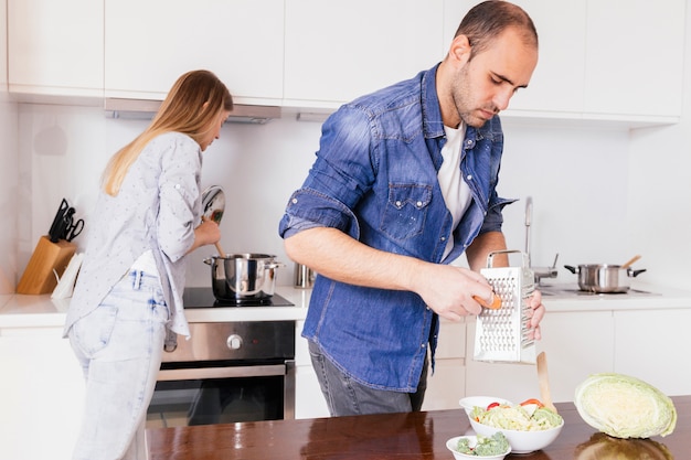 Zanahoria de rejilla del hombre joven con su esposa que prepara la comida en el fondo