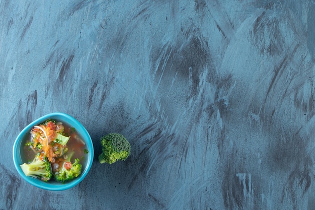 Zanahorias, brócoli y tazón de sopa de pollo, sobre fondo azul.